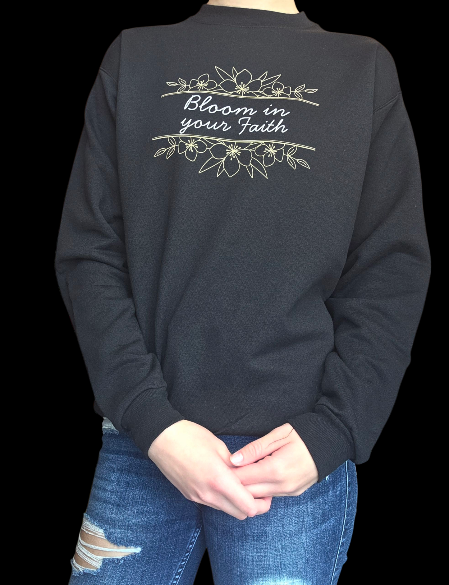 Black crewneck with 'Bloom in your Faith' text and floral design, worn by a person in blue jeans.