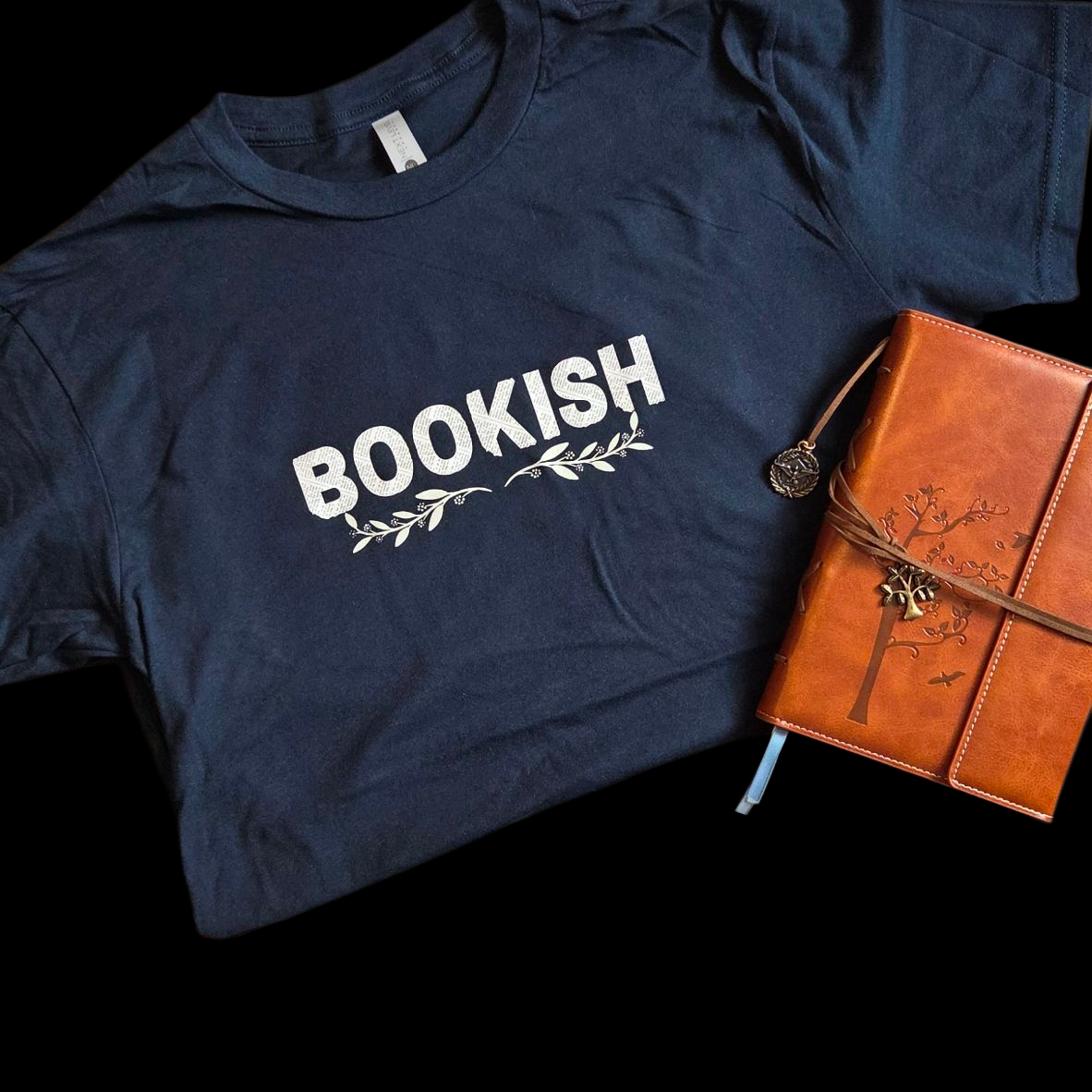 Navy blue t-shirt with 'BOOKISH' text and a leather-bound notebook on a black background